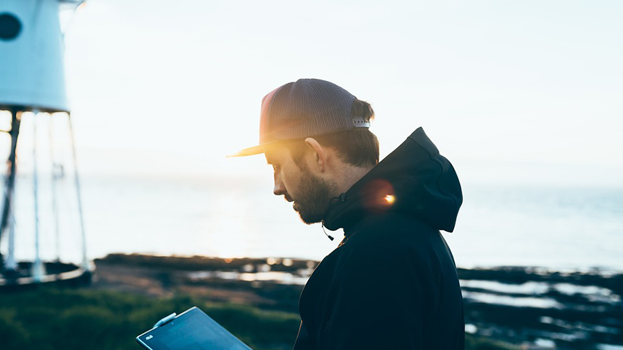 A man in a black jacket and cap stands near a coastal area at sunset, looking at a clipboard. A white structure is visible in the background, perhaps related to personnel management efforts on-site.