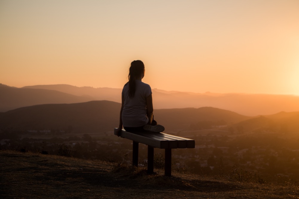 A person sits on a bench overlooking a mountainous landscape at sunset, finding solace and health and wellbeing support in the tranquil beauty of nature.