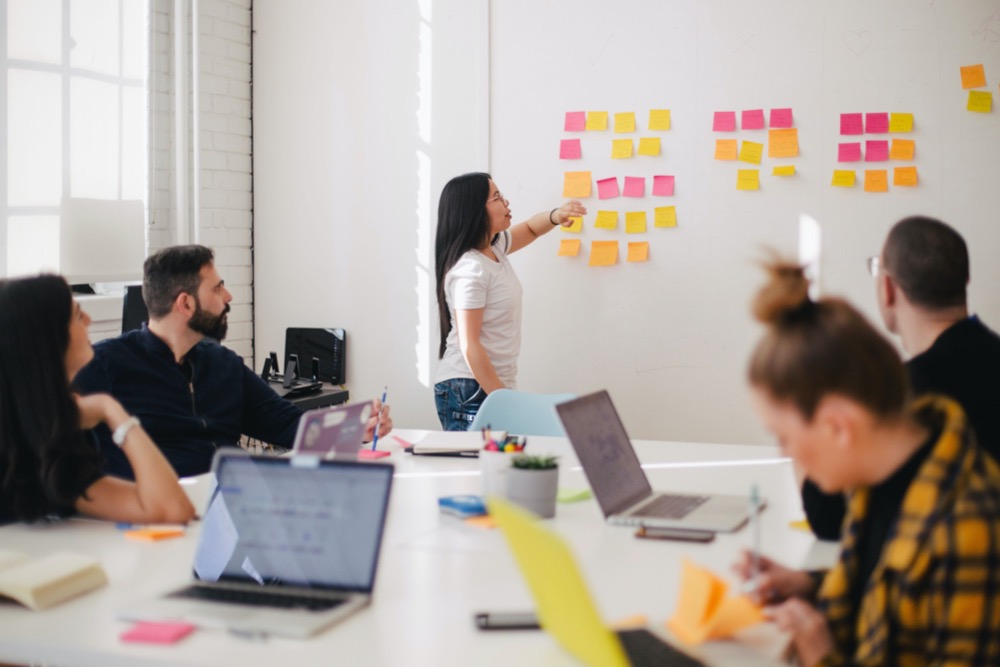 People in a meeting room with laptops, listening to a woman presenting ideas using sticky notes on a whiteboard during an employee training session.