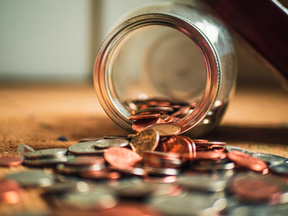 A glass jar tipped over, spilling assorted coins onto a wooden surface, a stark reminder of the cost of living crisis.
