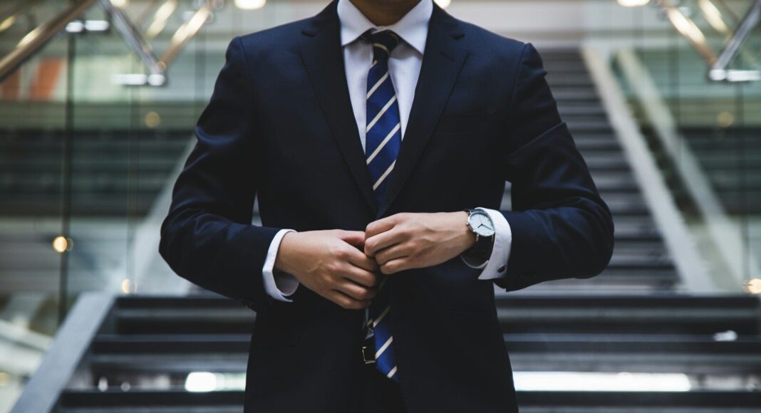 A man in a suit stands before the camera.
