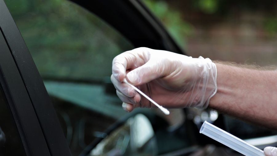 A gloved hand holding a swab near a car window, possibly for testing purposes, highlights the dedication despite NHS staff absences.