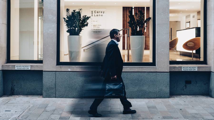 A man in a suit and tie walks briskly past a modern storefront with large planters, perhaps on his way to an important people management meeting. He carries a briefcase and appears blurred due to motion. The building sign reads 