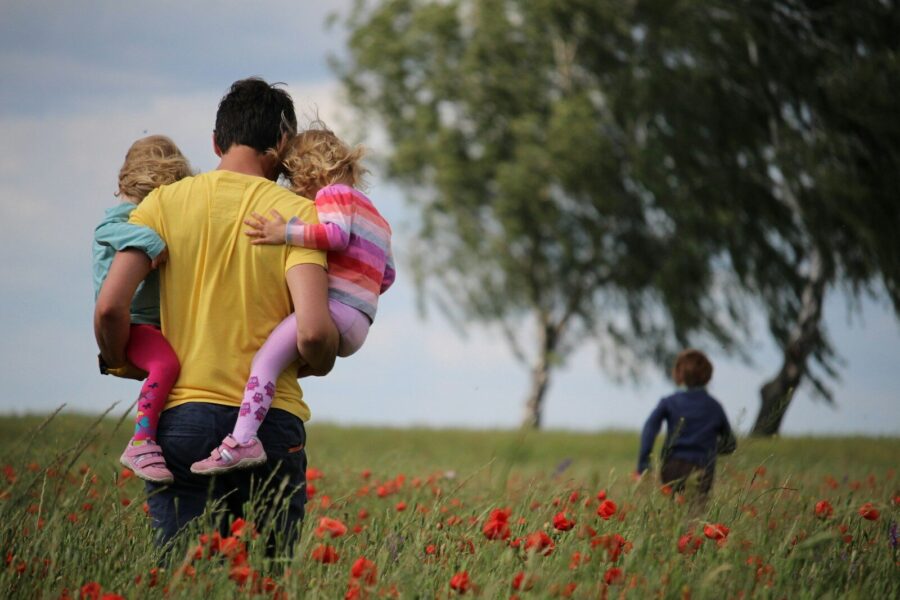 A man carrying two children in a field while another child runs ahead.