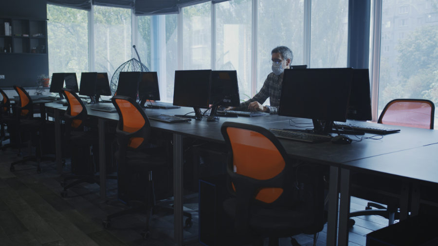 A person wearing a mask sits alone in a dimly lit office with several empty desks and orange chairs, facing multiple computer monitors. Large windows allow natural light to filter into the room, casting a somber mood as the news of Wales workers fined lingers in the air.