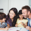 A family of four sits at a table. The parents engage with their two young children, who are holding pencils and appear to be working on a project or homework, mindful of the ever-present childcare costs in their lives.