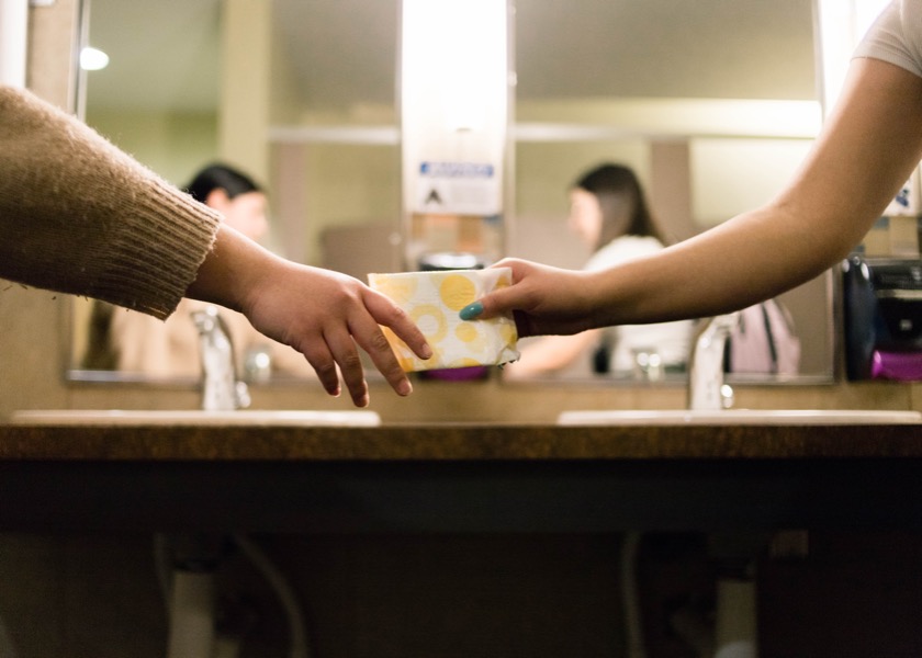 Two people exchanging a small yellow package over a bathroom counter, with sinks and mirrors in the background—a discreet moment that underscores the importance of women's health.