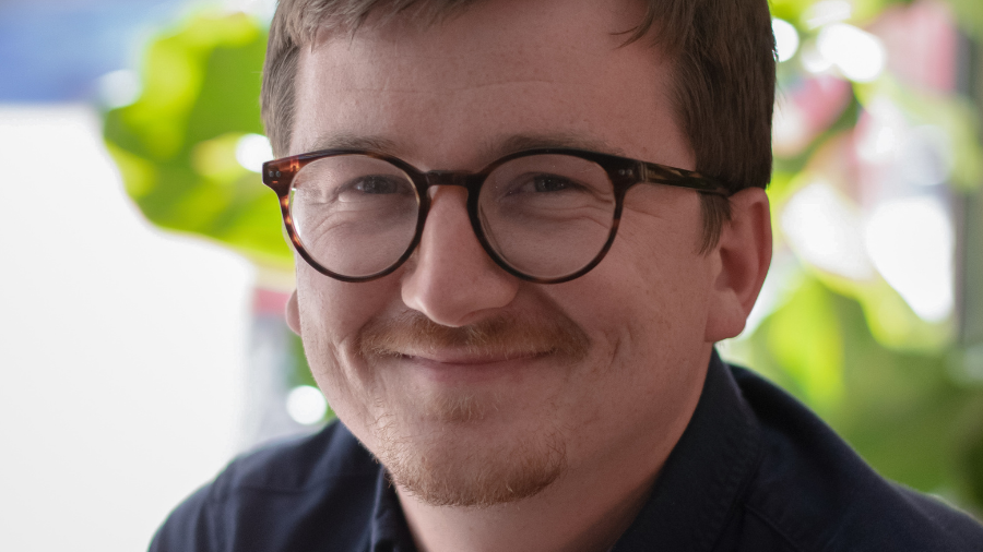 A person with short hair and glasses is smiling softly at the camera, exuding a sense of calm confidence. The background features blurred greenery, perhaps hinting at work-life balance or even thoughts of a secure workplace pension.