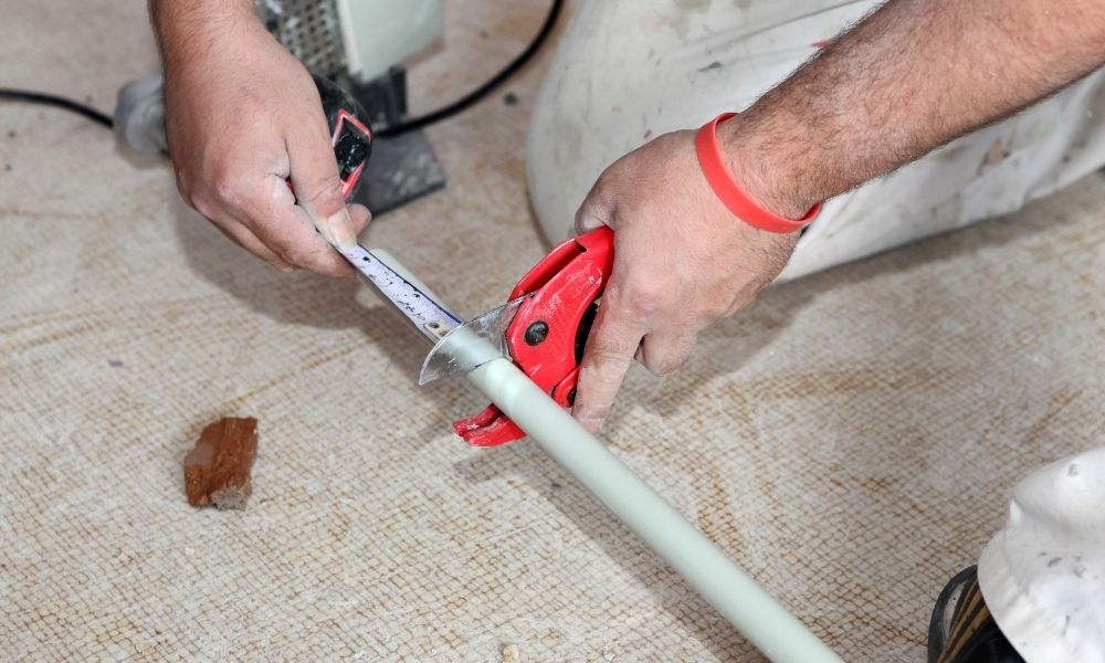 Hands using a red pipe cutter to cut a white PVC pipe while measuring with a tape measure, showcasing the precision of an engineer. A piece of wood and part of a machine are visible in the background.