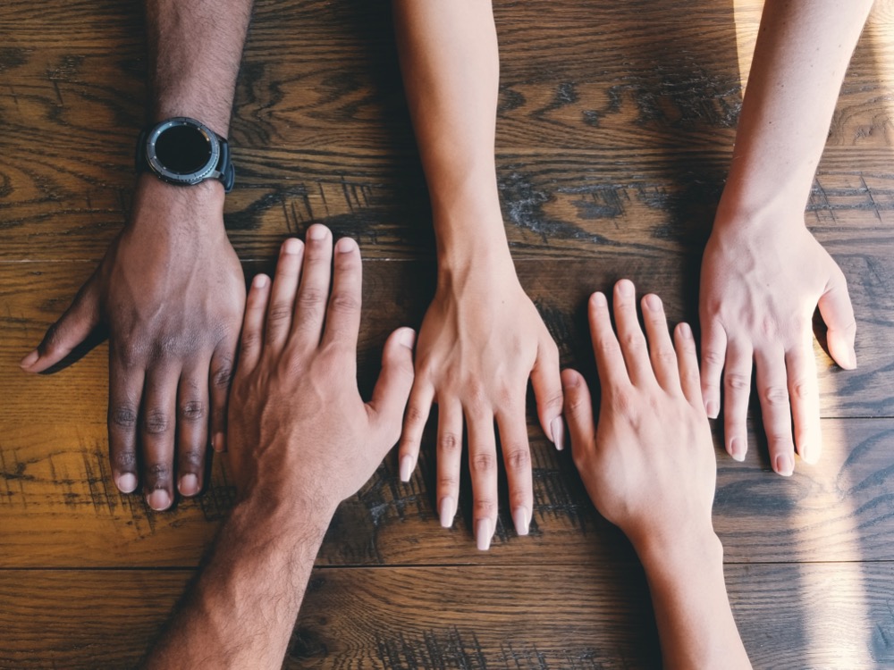 Four hands of different skin tones rest flat on a wooden table, with fingers extended and spread slightly apart, embodying the essence of diversity and inclusion.