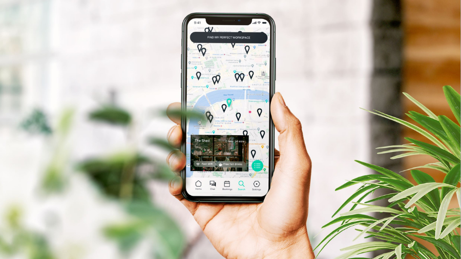 A person holding a smartphone displaying a map with multiple location pins marked, surrounded by indoor plants, highlighting a workspace designed to boost employee mental health.