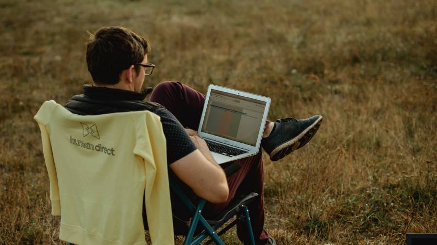A person is sitting outdoors on a blue folding chair, working on a laptop, presumably handling some HR tasks. They are wearing glasses and a black shirt, with a yellow jacket draped over the chair's back. The background is a grassy field.