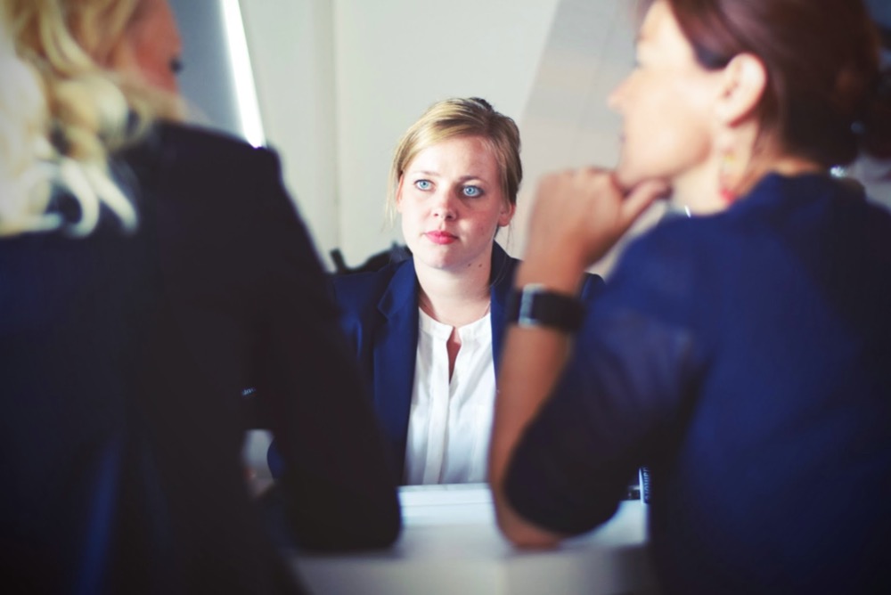 Three women in a business discussion sit around a table, one woman facing the camera while the other two have their backs turned. They appear to be in a professional setting, possibly addressing a resignation matter.