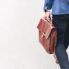 A person in a blue blazer and dark trousers walks on a gray pavement, holding a brown leather briefcase, possibly heading to a talent recruitment event.