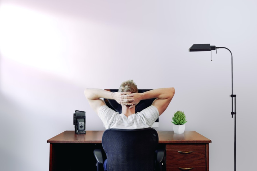 Person relaxing in an office chair with hands behind head, facing a computer on a wooden desk with a potted plant and black lamp nearby, embodying the ease of working from home.
