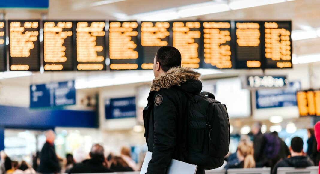 A male passenger stands in front of a departures board at an airport.