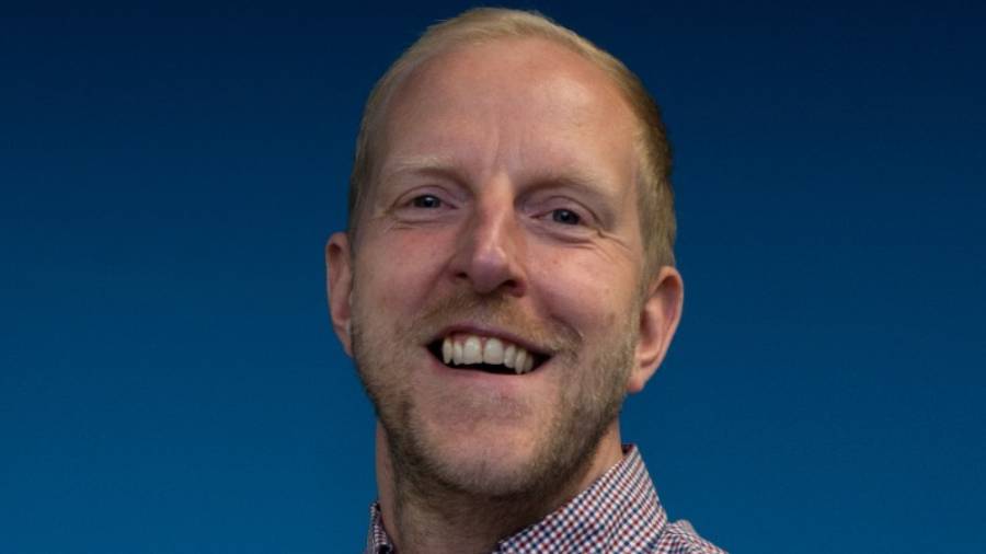 A man with short blonde hair and a beard smiles, wearing a checkered shirt as he poses against a dark blue background, embodying the essence of effective People Management.