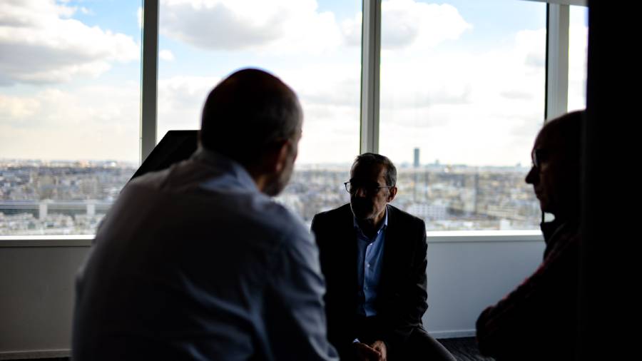 Three older workers are sitting and talking in a room with large windows. The cityscape is visible in the background through the windows, casting a serene backdrop as they discuss the recent furloughs.