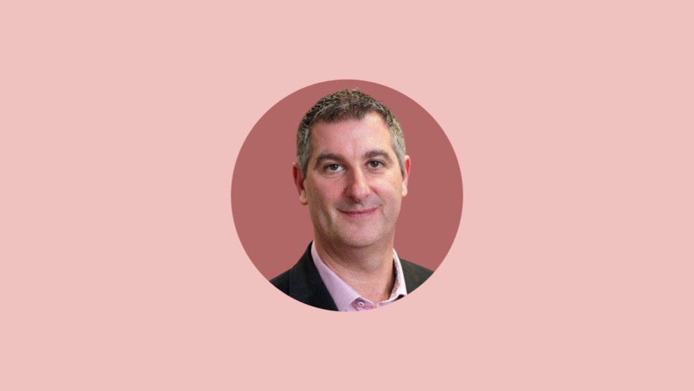 A portrait of a man with short gray hair, exuding quiet pride in his dark suit jacket over a light pink shirt, set against a pink background.