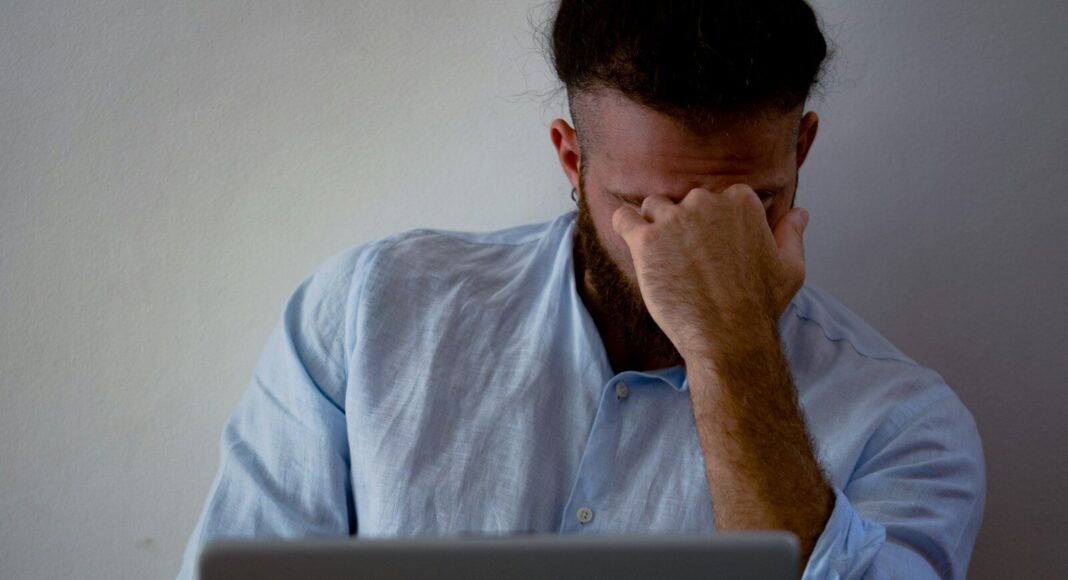 A man with his hand to his head while working at a laptop, suggesting illness at work.