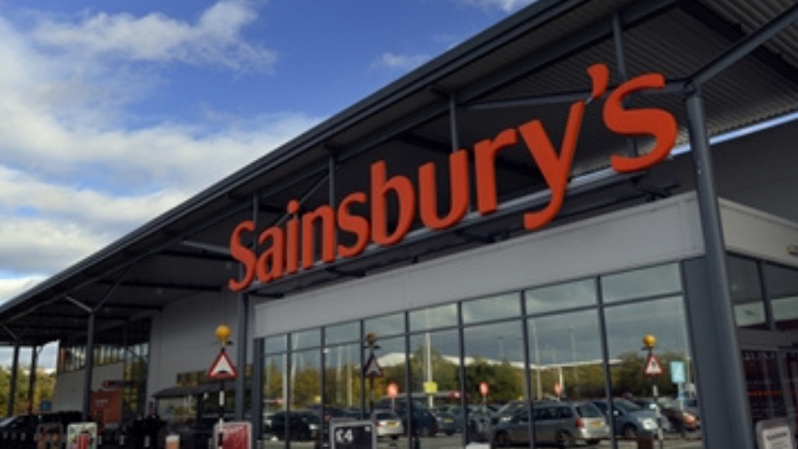 Exterior view of a Sainsbury's supermarket with the store's sign prominently displayed above the entrance and parked cars visible in the foreground. A banner proudly announces that Sainsbury's pays workers £10 an hour.