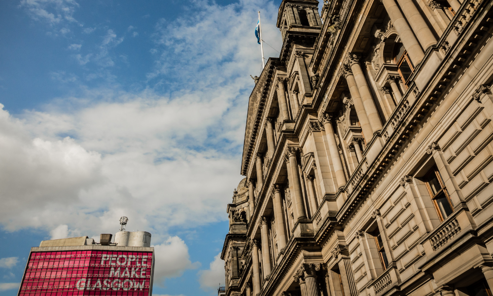 A close-up of a historic building against a blue sky with clouds. In the background, a modern building displays the message 
