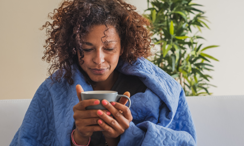A woman with curly hair, wrapped in a blue blanket, holds a mug with both hands while seated, contemplating her thoughts during Covid-19 isolation. A green plant in the background adds a touch of nature to her serene moment.