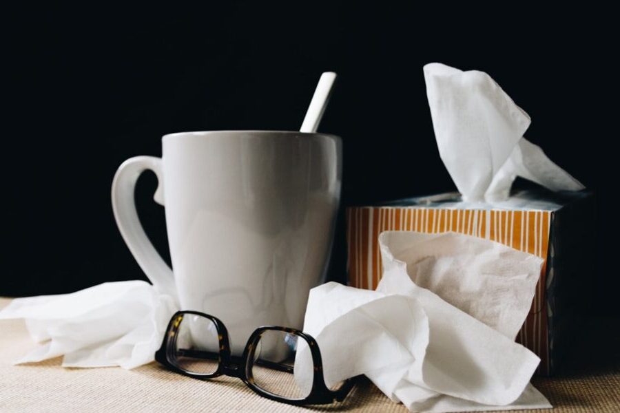 A white mug with a spoon, a few tissues, a box of tissues, and a pair of eyeglasses are placed on a beige surface against a black background, suggesting the essentials for someone on sick leave.