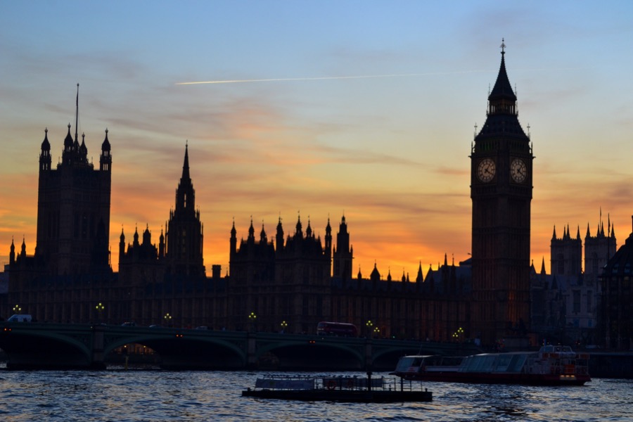 Silhouette of the Houses of Parliament and Big Ben at sunset, viewed from across the River Thames with a bridge and boats in the foreground, reminiscent of serenity before the bustling activity surrounding the Spring statement.