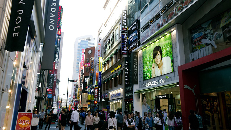 A bustling urban street lined with various shops and advertising signs. The street is crowded with people. One large display features a smiling person promoting 