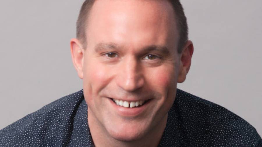 A person with short hair smiling at the camera against a plain background, wearing a dark blue polka-dotted shirt, embodies the essence of effective people management.