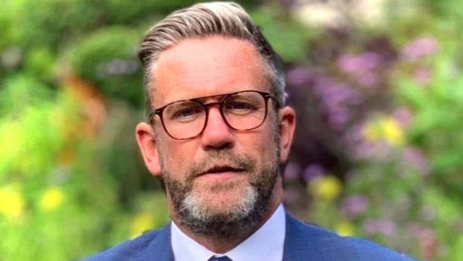 A man with glasses and a beard, wearing a suit, stands outdoors with a blurred background of greenery and flowers. His demeanor exudes the confidence of someone seasoned in Human Resources.