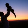 A man holds an infant up high at sunrise.
