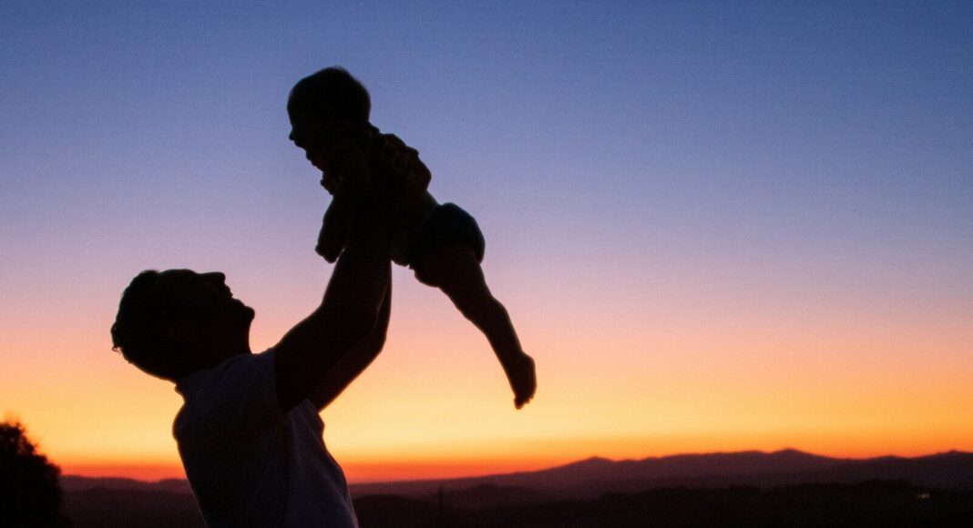 A man holds an infant up high at sunrise.