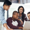 Four people gather around a laptop, collaborating and smiling in a brightly lit office setting, epitomizing the dynamic nature of tech jobs.