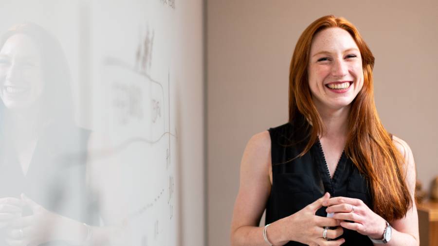A person with long red hair is smiling while standing indoors next to a whiteboard with HR diagrams.
