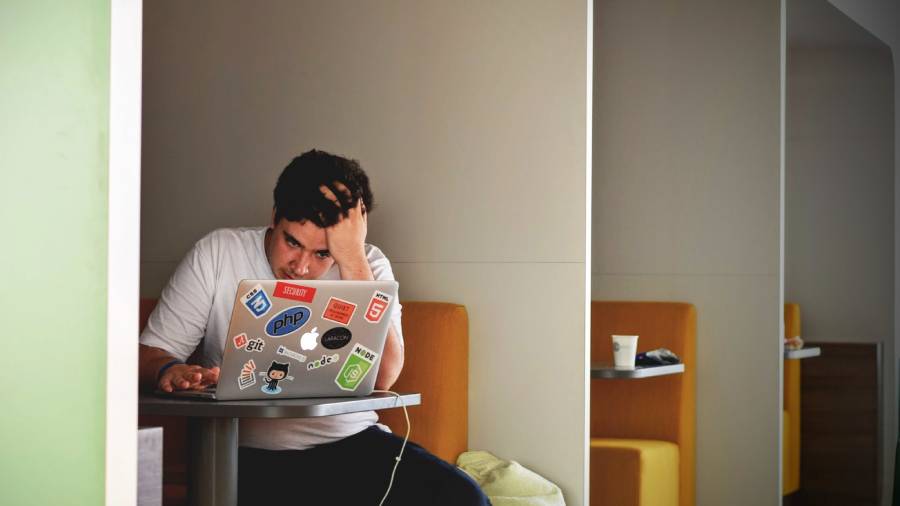 A person sits in a booth, leaning on one hand while using a laptop covered in stickers. They appear focused or stressed, possibly dealing with HR tasks or people management issues.