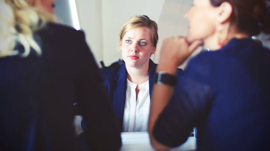Three women sit at a table having a conversation, focusing on the woman in the middle who, like an HR specialist, is looking at the others attentively.