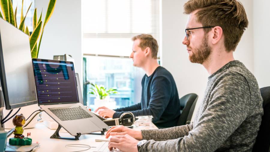 Two men working at desks with computers in a brightly lit, diverse office, one with a laptop on a stand and the other with a desktop monitor.