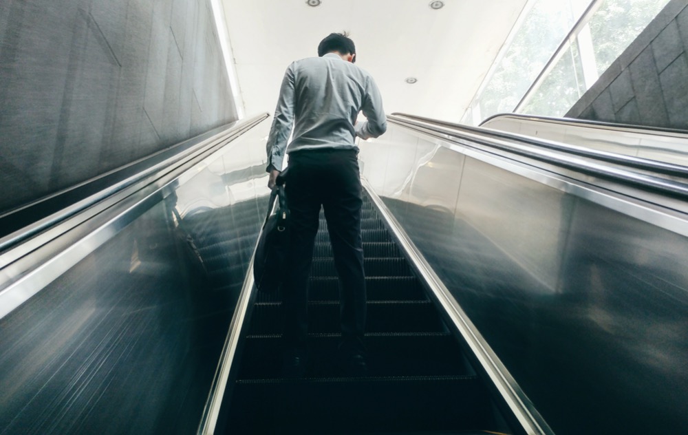 A man in a light shirt and dark pants ascends an escalator, holding a bag in his left hand, visible from behind. His expression reflects the weight of the rising cost-of-living.