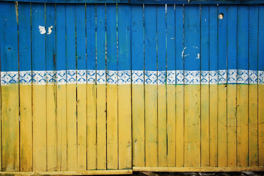 A wooden fence is painted with blue on the upper half and yellow on the lower half, featuring a white decorative pattern in the middle, symbolizing solidarity to help Ukrainian refugees.