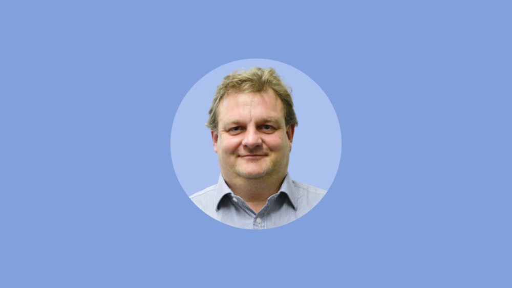 A man with light brown hair is smiling at the camera, wearing a light blue shirt, embodying the ease of hybrid working. The background is solid light blue.