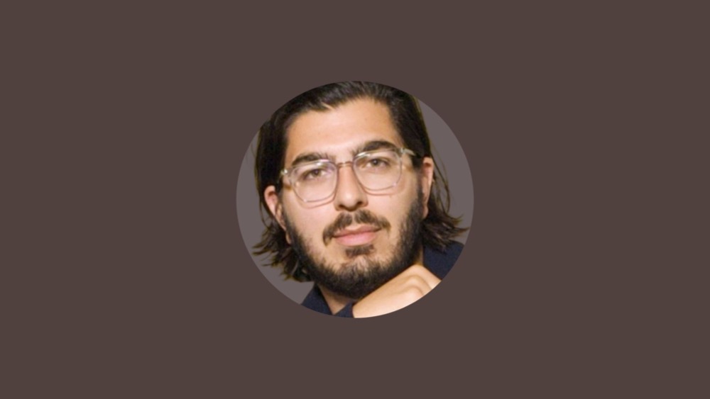 A person with shoulder-length hair, a beard, and glasses, looks at the camera against a plain background, embodying the professional yet approachable vibe you’d expect from someone well-versed in employee benefits.