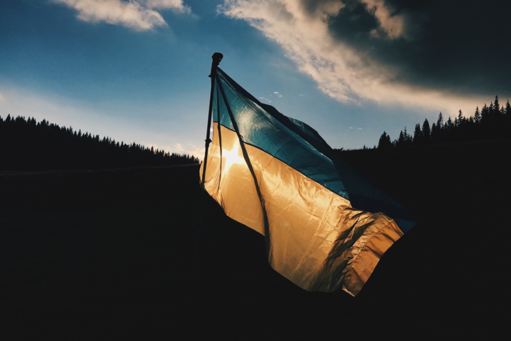 A Ukrainian flag waves in the wind against a backdrop of silhouettes of trees and a dramatic sky during sunset, symbolizing resilience amid tensions with Russia.