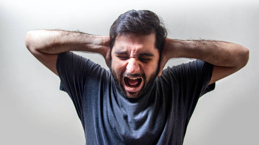 A bearded man in a dark shirt closes his eyes and screams, holding his hands over his ears against a plain background, evoking the stresses often faced in HR.