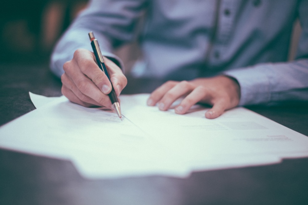 A person in a blue shirt writes on white paper documents with a black and gold pen, preparing for the jobs market.