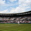 A wide shot of a tennis match being played on a grass court in a large, open-air stadium filled with spectators under a partly cloudy sky, capturing the essence of sporting success.