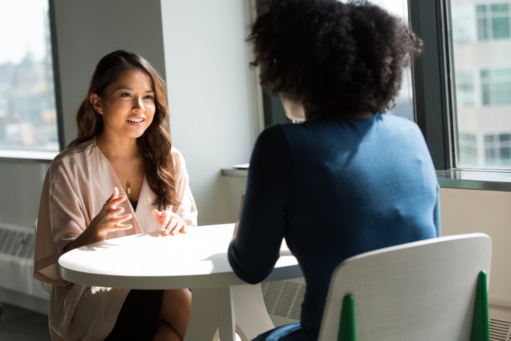 Two individuals are seated at a small round table in an office setting, engaged in a conversation. One person is facing the camera and gesturing while speaking, likely discussing a pay rise, and the other has their back to the camera.