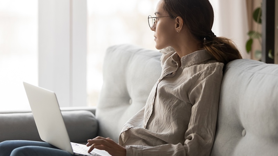 A person with glasses sits on a couch, looking to the side while using a laptop on their lap, perhaps working on some Human Resources tasks.