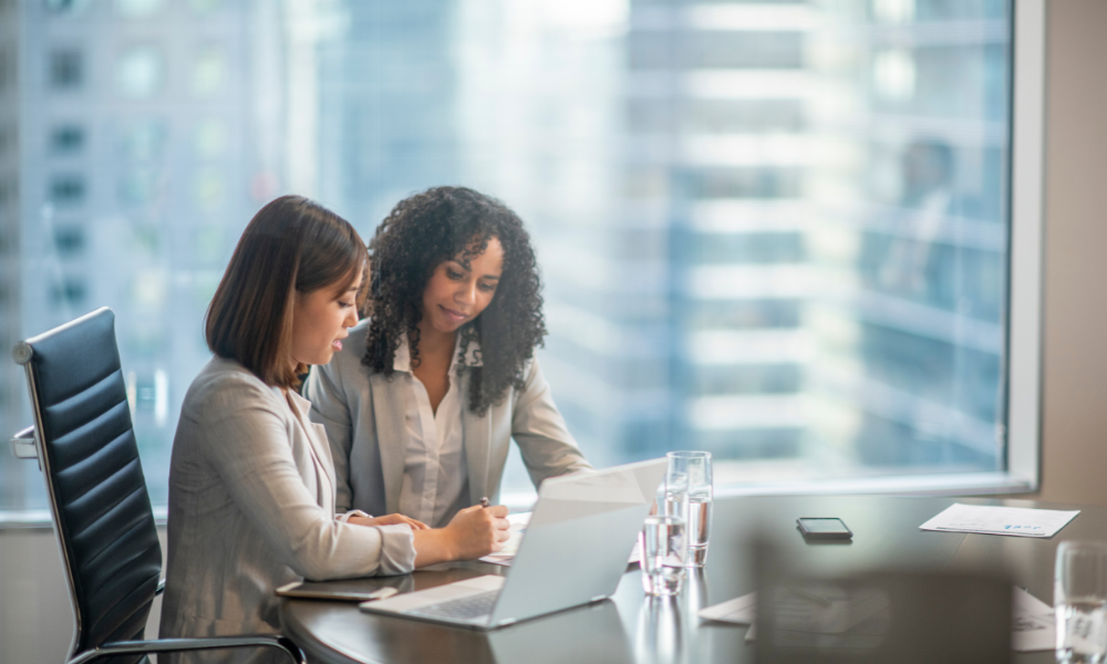 Two women sit at a conference table in an office setting, working on a laptop and reviewing documents. Large windows in the background reveal a cityscape with tall buildings, as they discuss balancing career goals with the challenges of parenthood.